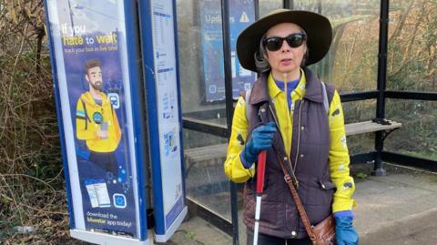 Patricia Clements is standing at a bus stop. She is facing towards the camera, in front of the bus shelter, which is surrounded by sparce hedging and trees and stands next to a sign which has all the bus timetables on it. Patricia has shoulder length grey hair and wears a wide brimmed forest green hat and black plastic framed square sunglasses. She has a yellow blouse on, which has a print of illustrated greyhounds on it, with a blue top on underneath and a padded brown collared gilet, which is zipped up. Patricia has a tan brown leather cross body bag on, blue leather gloves and is holding her cane, which has a red handle and is white further down.