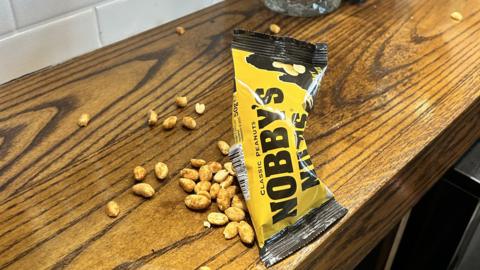 An opened packet of dry roasted Nobby's Nuts sits in the centre of the image, with several nuts strewn across a wooden counter. The packet is bright yellow with black text on it