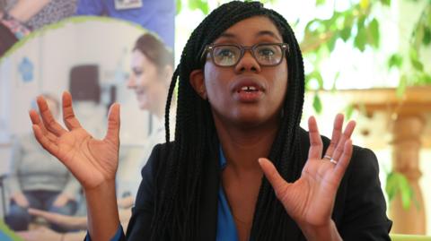 Kemi Badenoch holding her hands in the air. She is wearing a black blazer over a blue top, and has her mouth open while speaking in a sitting room at a hospice.