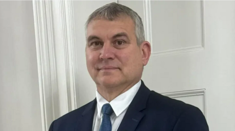 A man with short grey hair is looking into the camera and smiling. He is wearing a white shirt and blue tie and a blue jacket. He is standing in front of a white door.