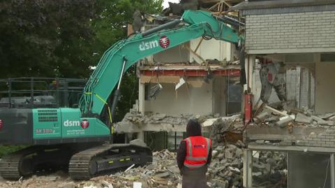 A green bulldozer demolishing a white brick office block. A worker in a high visibility looks on. 