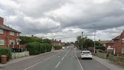A straight residential road running through a housing estate. The road has two lanes marked with dashed white centre lines and wide pavements on both sides. Several cars are parked along the street, including a white car parked on the right-hand side in the foreground and additional vehicles further down the road.
On the left side of the image, there is a row of two-storey red-brick terraced or semi-detached houses with dark grey tiled roofs and brick chimneys. The houses have white-framed windows and small front gardens enclosed by hedges, low fencing, and gates. Wheelie bins are visible near one of the properties, and a brown litter bin stands beside the pavement.