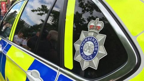 Close up of the side of a Humberside Police patrol car with the force's badge in the rear side window and bright yellow and blue police markings.