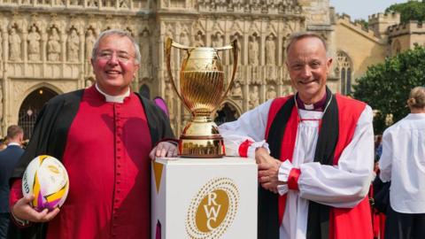 Two people wearing red and black religious robes stand beside a gold trophy. The man on the left is holding a rugby ball. There is a cathedral in the background.