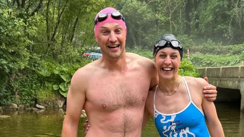Brother and sister Hugh and Alex Addison are standing in a river with a concrete bridge behind them, wearing swimming costumes and smiling broadly. He has a pink swimming cap and she has a black one. Both have swimming goggles resting on the top of their heads.