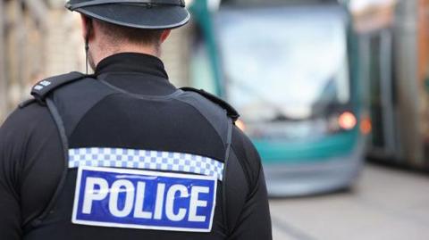 A police officer stands with his back to the camera. He is wearing a black uniform which includes a vest with the word "police" written on it. A Nottingham tram can be seen in the background.