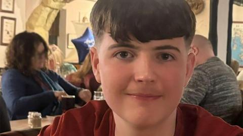Head-and-shoulders photograph of a smiling Liam Bracken sitting in a restaurant. He has dark hair, longer on top and cropped at the sides and is wearing a burgundy t-shirt
