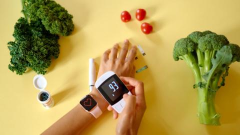 A person check their glucose level on a small monitor. The background is yellow and there are various fruits and veg scattered around.