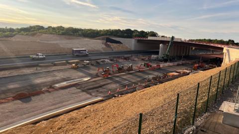 A shot of the A417 Missing Link project in Gloucestershire. It is a newly built road and traffic cones and diggers can be seen on the roadside. There is a bridge over the road.