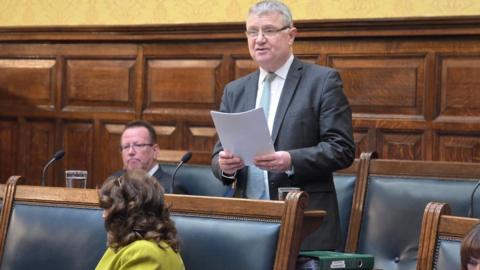 Chris Thomas, a man with grey hair and glasses reads from a piece of paper, he wears a suit in the Tynwald Chamber.