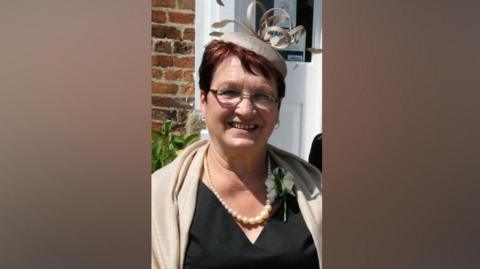 A woman is smiling while standing in front of the door of a house. She is dressed in formal wear, with a black top or dress, cream-coloured pashmina, a white or cream-coloured rose buttonhole, pearl necklace and a cream coloured pillbox hat with fascinator detail. She is also wearing glasses and she has red-brown short hair.