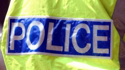 The back of a police officer wearing a yellow hi-vis with police in white capital letters written on a blue background