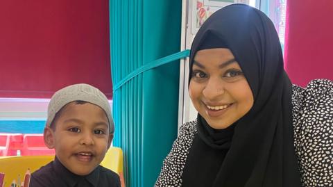 A boy in a grey hat and a stripy navy jumper sits beside a lady in a black and white dress and a black hijab. They are both holding an orange pencil and writing. They are in a classroom sitting at a yellow table with yellow seats and red blinds behind them