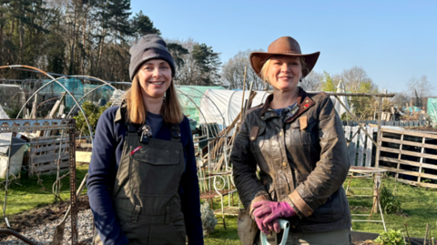 Two smiling women stood in an allotment holding gardening equipment 