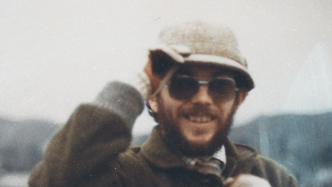 Kenner Elias Jones is a white man in his 30s or 40s in this picture, with a brown beard and moustache. He wears dark lensed prescription glasses and a cream coloured hat which he is holding, a checked shirt and tie covered by a brown coat with a collar. He is smiling at the camera. The image is grainy.