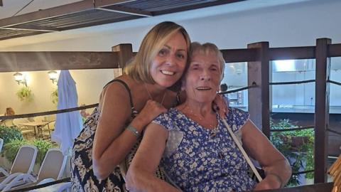 Tracey Davison and mother Mary Curtis taking a holiday photo. They are in the outdoor area of a restaurant. It it summer and they both wearing dresses. There are lights on in the background and sun loungers. Curtis is sitting down and has grey hair. Davison is smiling and putting her arms on Curtis's shoulders. She has blond hair and is wearing a black and white patterned dress, whereas Curtis is wearing a blue and white patterned dress.