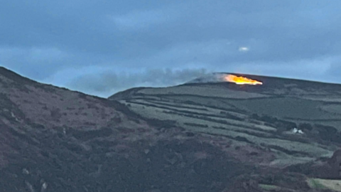 A fire seen in the distance on a patch on the hillside, with grey skies behind.