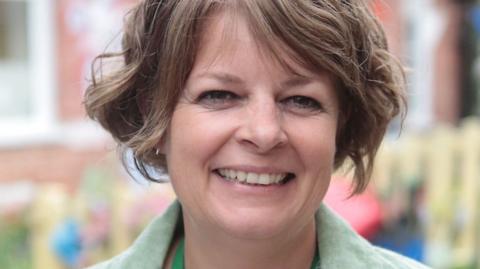 A headshot of Ruth Perry, head teacher of Caversham Primary School. She is smiling at the camera and wearing a green jacket. Her hair is brown, cropped and worn in curls