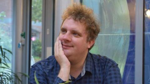 John Osborne, who has curly ginger/brown hair, is smiling and looking thoughtfully away from the camera. He is seated and wearing a blue checked shirt. His right hand is partly across his face.