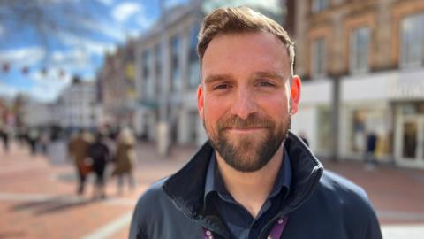 William Russell is wearing a blue shirt and blue jumper. He has a brown beard and short, brown hair.