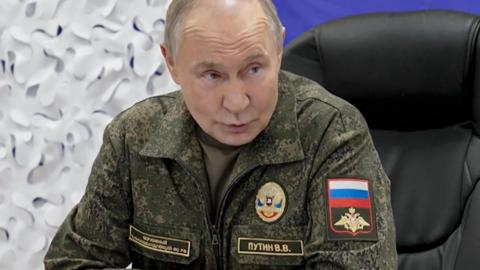 Russian President Vladimir Putin speaks during a briefing to journalists at a military base. He is wearing a camouflage uniform and sat in a black office chair. The background of the image is a blue and white backdrop.