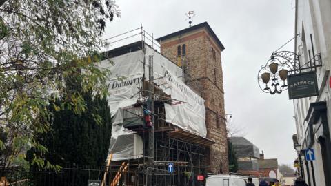 A church covered in scaffolding on the left of the picture. A white curtain has been around the scaffolding. On the right side, in the foreground, are shops, including FatFace.