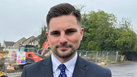 Joe Harris, a man in his 40s with short brown hair, a suit jacket on, a white shirt and a blue tie with white polka dots looks directly at the camera as he stands in front of a building site in the rain.