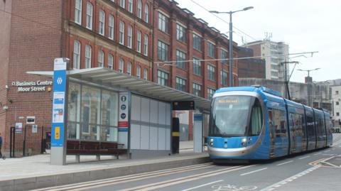 A blue and silver tram pulling up to a tram stop on a city street by a four-storey building with white lettering on the wall saying Business Centre Moor Street.