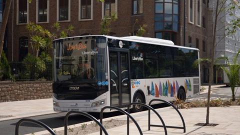 A white LibertyBus bus travels along a paved city street bordered by landscaped areas and modern buildings. Bicycle stands line the foreground, while palm-like trees and a stone wall sit beside the road.