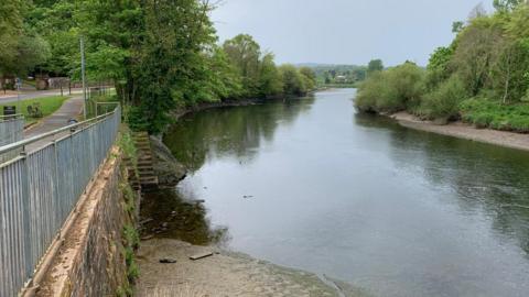 A winding stretch of the River Nith with gravelly banks and trees down either side with hills in the distance and hazy, cloudy, light blue sky overhead. There is a footpath on the left hand side of the river, and steps can be seen going down into the water. 