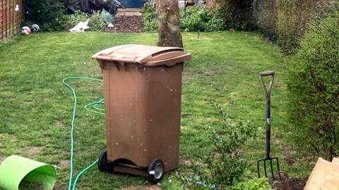 A brown garden waste bin sits in a garden. A green hose pipe lies on the grass around it as well as a garden fork.