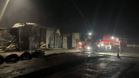 A night-time scene showing a corrugated steel walled building damaged by fire.