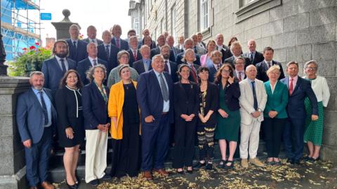 Members of the States of Guernsey. All 40 of them, a mix of men and women in business attire, are standing on the steps of the Royal Court. 
