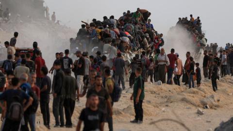 A group of people walking on sandy, rocky ground. A large group of people appear to have climbed onto two trucks, one in the centre of the image and one in the background.