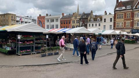 Cambridge Market Square, which has trading stalls under canopies, with a flower stall closest to the camera. Members of the public are walking around the market.