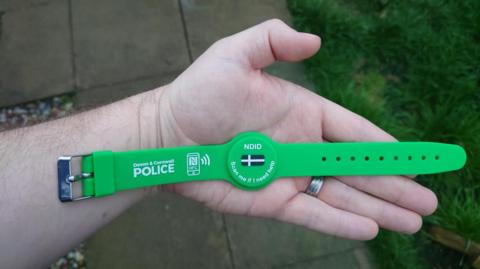 A hand laying flat. There is a wristband lying on the palm of the hand. It is green, has a Cornish flag in the centre and says "NDID" and "Scan me if I need help" in the centre. On the left strap it says "Devon and Cornwall Police" with a picture of a phone next to it. 