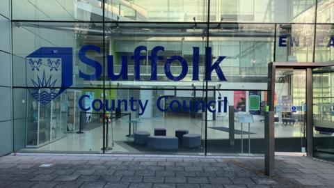 A glass-fronted building which is the headquarters of Suffolk County Council. It reads Suffolk County Council in large blue writing