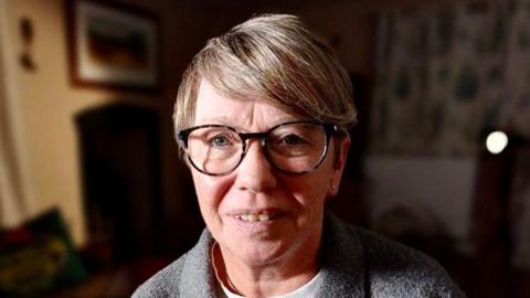 Amanda McKay, in her late 50s, with glasses and a grey top, with a dark background  