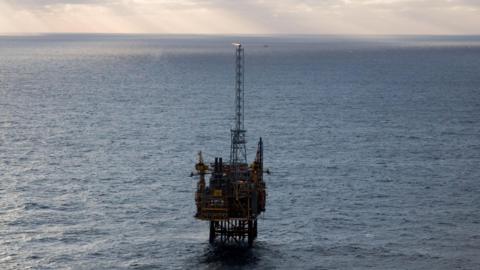 Offshore oil platform standing alone in the sea under a cloudy sky, with rays of sunlight breaking through the clouds and a small flare burning at the top of the structure