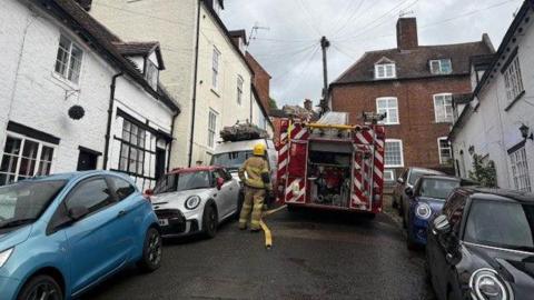A fire engine is parked on a steep, narrow street, with two sets of cars either side of it. There is also a firefighter in full gear and a hose on the floor. There are terraced cottages on either side of the street.