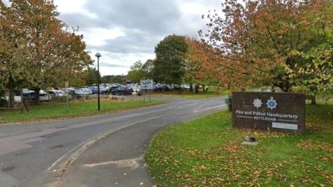 Shot of the entrance to the police headquarters in Nettleham. There is a sign which reads 'Fire and Police Headquarters', along with a sign denoting the different areas of the car park further away. The road is marked with double yellow lines.