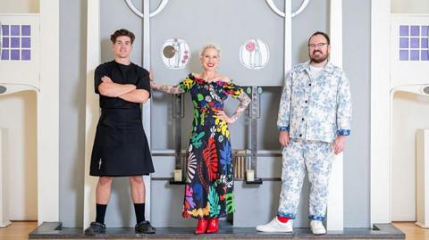 Danny Campbell, Anna Campbell-Jones and Banjo Beale stand in a line in front of a beautiful fireplace inside the House for an Art Lover, where the winner was announced. Danny wears a black t-shirt, black kilt and Crocs, Anna, with her bright white cropped hair, wears a bold floral dress and red boots and Banjo rocks a blue and white china pattern-style suit with white trainers.
