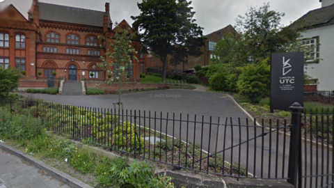 A Google Street View image of Lincoln University Technical College, an old red brick building with a landscaped green area to the front, behind a black metal fence.