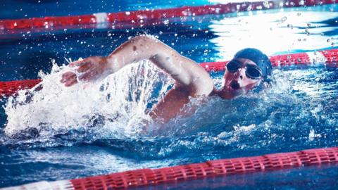 A stock image of a man wearing goggles and a swimming cap swimming in a pool.