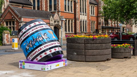 A big blue and patterned barrel is positioned in the town next to a floral display. It is tilted backwards and resting on a square base.