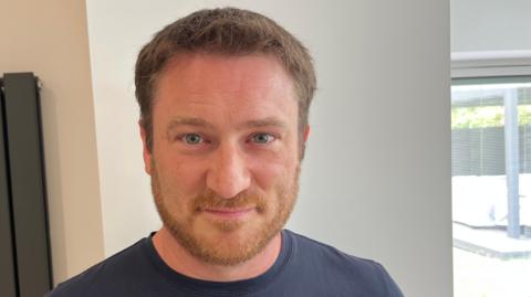 Brett Rigby has short reddish hair and matching beard and moustache, he has a blue top on and behind him is a living room wall with a grey vertical radiator.