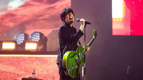 Texas singer, Sharleen Spiteri, in a black glittery suit, holds a green guitar and sings into a microphone. She has short dark hair and is surrounded by stage lights.