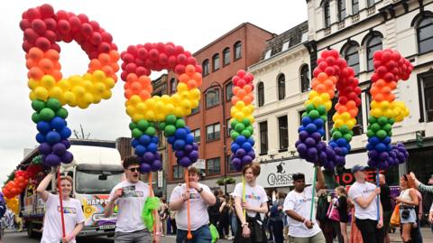People taking part in the Belfast Pride parade in the city centre in 2024 on the street, they are wearing white  t-shirts are holding balloons that read PRIDE in red, orange, yellow, green and blue. They are walking down a Belfast street with crowds watching on. 