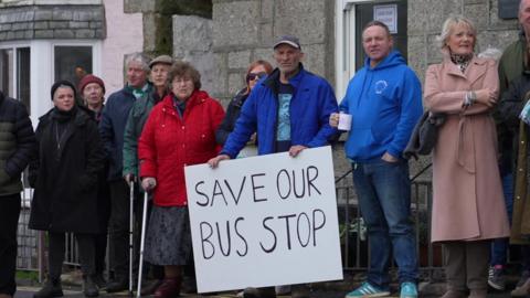 A group of nine people wearing winter clothes standing in front  of a building holding a large white sign. The sign reads 'Save Our Bus Stop'