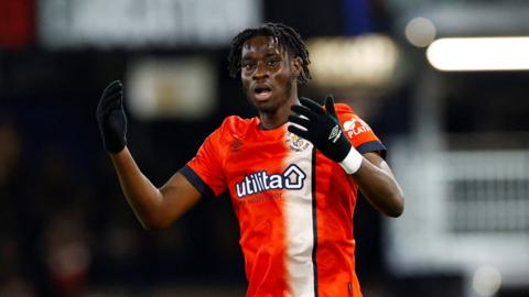 Elijah Adebayo has his mouth agape and throws his hands out in front of him in annoyance. He wears an orange luton football shirt and black gloves